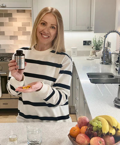 Woman holding a biocuria supplement bottle and capsules in a kitchen with fruits on the counter.