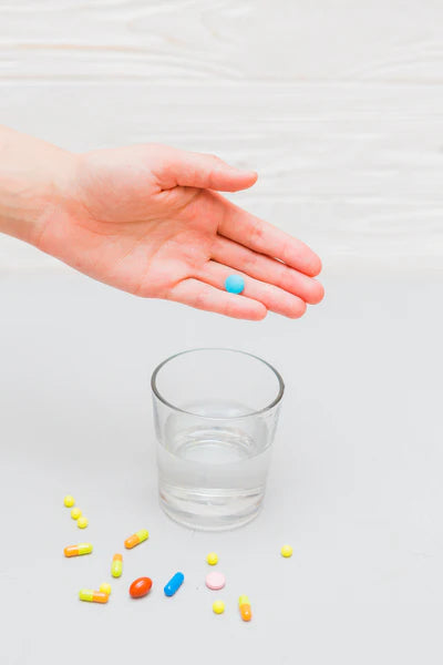 Hand holding a blue pill with a glass of water and scattered pills on a light surface